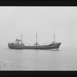 Schwarzweißaufnahme eines Frachtschiffs in Seitenansicht, das ruhig über das Meer fährt; das Bild zeigt einen breiten Filmrand und ein Wasserzeichen des Internationalen Maritimen Museums Hamburg in der unteren linken Ecke.