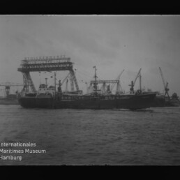 Schwarzweißaufnahme eines Frachtschiffs, das im Hafen vor einem großen Portalkran und mehreren Ladekränen liegt, mit Wasser im Vordergrund und dem Logo ‚Internationales Maritimes Museum Hamburg‘ unten links.