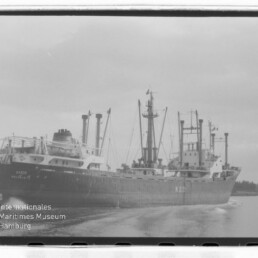 Schwarzweißfoto eines großen Frachtschiffs mit mehreren Masten und Ladekränen, das auf einem Kanal fährt und eine Bugwelle zieht; links unten Wasserzeichen des Internationalen Maritimen Museums Hamburg.