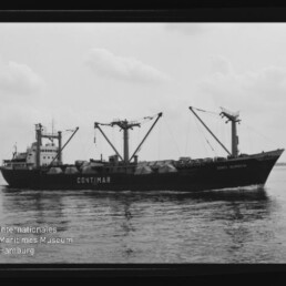 Schwarzweißaufnahme eines Frachtschiffs mit der Aufschrift CONTIMAR, mehreren Ladebäumen und planenbedeckter Decksladung, auf ruhiger See unter bedecktem Himmel; Wasserzeichen des Internationalen Maritimen Museums Hamburg unten links.