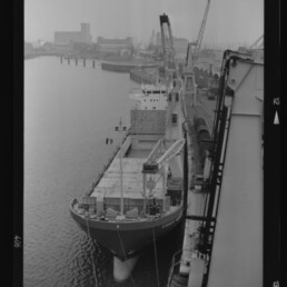 Schwarzweiß-Aufnahme eines am Industriekais liegenden Frachtschiffs aus erhöhter Perspektive, an Bord und am Kai mehrere Hafenkräne beim Verladen, im Hintergrund Hafenanlagen und Silos.
