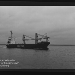 Frachtschiff CONTIMAR mit zwei Ladekränen fährt bei ruhigem Meer unter bewölktem Himmel vor einer niedrigen Küstenlinie; Museumswasserzeichen unten links.