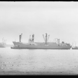 Schwarzweißaufnahme eines Frachtschiffs mit mehreren Ladekränen, das in einem Hafen auf ruhigem Wasser liegt; im Hintergrund weitere Schiffe und Hafenkräne, unten links Wasserzeichen des Internationalen Maritimen Museums Hamburg.