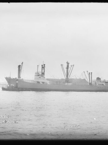 Schwarzweißaufnahme eines Frachtschiffs mit mehreren Ladekränen, das in einem Hafen auf ruhigem Wasser liegt; im Hintergrund weitere Schiffe und Hafenkräne, unten links Wasserzeichen des Internationalen Maritimen Museums Hamburg.