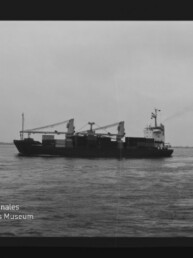 Schwarzweiß-Aufnahme eines seitlich sichtbaren Frachtschiffs mit zwei Ladekränen und leichter Rauchfahne auf ruhiger See; schwarzer Filmrand und Wasserzeichen des Internationalen Maritimen Museums Hamburg unten links.