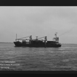 Schwarzweiß-Aufnahme eines seitlich sichtbaren Frachtschiffs mit zwei Ladekränen und leichter Rauchfahne auf ruhiger See; schwarzer Filmrand und Wasserzeichen des Internationalen Maritimen Museums Hamburg unten links.