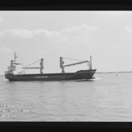 Schwarzweiß-Fotografie eines Frachtschiffs mit großem Schriftzug CONTIMAR und zwei Ladekränen auf dem Deck, das bei ruhiger See unter leicht bewölktem Himmel entlangfährt; Negativrand des Films und Wasserzeichen des Internationalen Maritimen Museums Hamburg sichtbar.