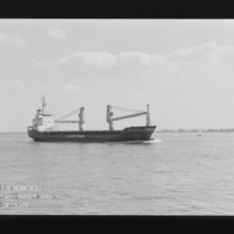 Schwarzweißaufnahme eines Frachtschiffs mit der Aufschrift CONTIMAR und zwei Ladekränen, das auf ruhiger See vor flachem Horizont mit vereinzelten Booten fährt; Filmnegativrand und Wasserzeichen des Internationalen Maritimen Museums Hamburg sichtbar.