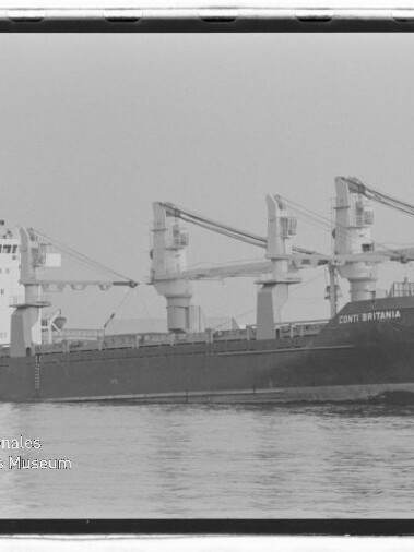 Schwarzweiß-Foto eines Frachtschiffs mit drei sichtbaren Ladekränen und dem Namen CONTI BRITANNIA am Bug, das ruhig im Wasser vor einem Hafen liegt; links unten Wasserzeichen des Internationalen Maritimen Museums Hamburg.