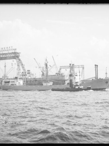 Schwarzweißaufnahme: Blick über die bewegte Hafenfläche auf mehrere Frachtschiffe und einen Schlepper im Vordergrund, dahinter massive Werftkräne und ein Kranlaufgerüst mit Firmenaufschrift.