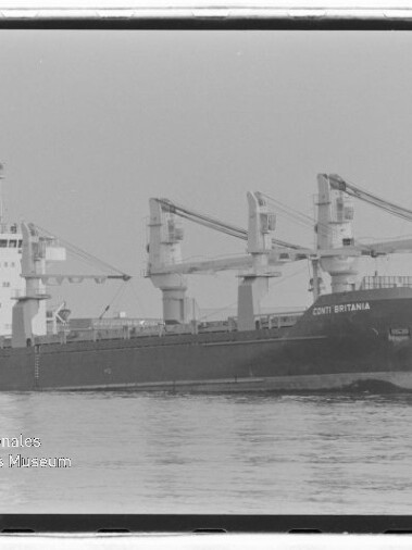 Seitenansicht des Frachtschiffs Conti Britannia mit drei Ladekränen, auf dem Wasser im Hafen vor Lagergebäuden.
