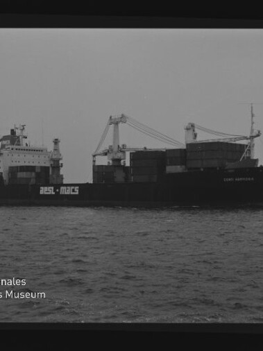 Schwarzweißaufnahme eines fahrenden Containerschiffs mit mehreren Deckskränen und gestapelten Containern auf ruhigem Meer, flachem Horizont und Museumswasserzeichen unten links.