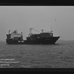 Schwarzweißaufnahme eines Containerschiffs mit aufgetürmten Containerstapeln und Ladekränen, das auf ruhiger See vor flachem Horizont fährt; unten links Wasserzeichen des Internationalen Maritimen Museums Hamburg.
