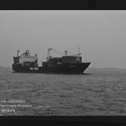 Schwarzweißaufnahme eines Containerschiffs mit aufgetürmten Containern und Ladekränen auf ruhiger See; sichtbarer Filmrand mit Aufschrift AGFA PAN 100 und Wasserzeichen des Internationalen Maritimen Museums Hamburg.