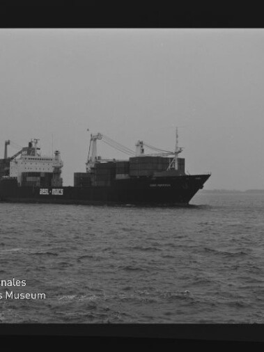 Schwarzweißaufnahme eines Containerschiffs mit aufgetürmten Containern und Ladekränen auf ruhiger See; sichtbarer Filmrand mit Aufschrift AGFA PAN 100 und Wasserzeichen des Internationalen Maritimen Museums Hamburg.