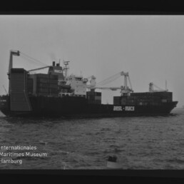 Schwarz-weiß-Aufnahme eines großen Containerschiffs mit aufgestapelten Ladecontainern und zwei Ladekränen, seitliche Ansicht auf dem Meer; im Vordergrund eine Boje und unten links das Wasserzeichen des Internationalen Maritimen Museums Hamburg.