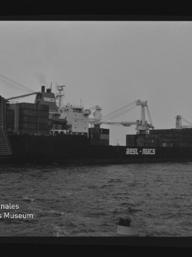 Schwarz-weiß-Aufnahme eines großen Containerschiffs mit aufgestapelten Ladecontainern und zwei Ladekränen, seitliche Ansicht auf dem Meer; im Vordergrund eine Boje und unten links das Wasserzeichen des Internationalen Maritimen Museums Hamburg.