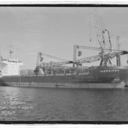 Schwarzweißaufnahme eines Frachtschiffs namens THORSIMBA mit aufgestellten Ladekränen am Kai, im Vordergrund Wasser und dem Wasserzeichen des Internationalen Maritimen Museums Hamburg