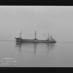 Schwarzweißaufnahme eines Frachtschiffs mit der Aufschrift CONMAR auf ruhiger See, seitliche Ansicht mit Ladekränen an Deck, Spiegelung im Wasser und sichtbarem Filmrand mit Museumslogo.