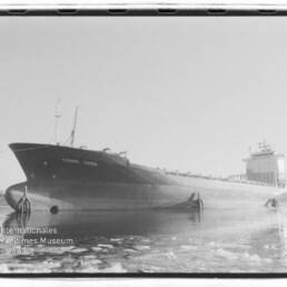 Schwarzweißfoto eines großen Frachtschiffs namens Conti Jork, seitlich im Hafen auf teilweise zugefrorenem Wasser mit Eisschollen liegend, mit markantem Bug und Aufbauten achtern; Wasserzeichen des Internationalen Maritimen Museums Hamburg unten links.