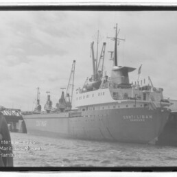 Schwarzweißaufnahme des Frachtschiffs Conti Liban (Hamburg) am Kai mit mehreren Ladekränen an Deck, Hafenbecken im Vordergrund, Lagergebäuden im Hintergrund und Wasserzeichen des Internationalen Maritimen Museums Hamburg unten links.