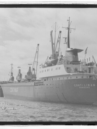 Schwarzweißaufnahme des Frachtschiffs Conti Liban (Hamburg) am Kai mit mehreren Ladekränen an Deck, Hafenbecken im Vordergrund, Lagergebäuden im Hintergrund und Wasserzeichen des Internationalen Maritimen Museums Hamburg unten links.