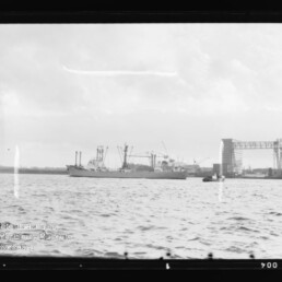 Schwarzweiß-Foto eines Frachtschiffs auf dem Wasser vor einem Werftgelände mit Kränen und einem großen Portalkran beschriftet DEUTSCHE WERFT, im Vordergrund welliges Wasser und bewölkter Himmel.