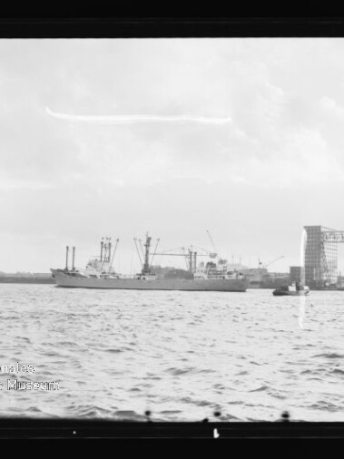 Schwarzweiß-Foto eines Frachtschiffs auf dem Wasser vor einem Werftgelände mit Kränen und einem großen Portalkran beschriftet DEUTSCHE WERFT, im Vordergrund welliges Wasser und bewölkter Himmel.