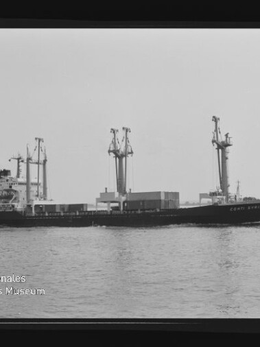 Schwarzweißfoto eines Frachtschiffs mit drei Ladekränen und Containern an Deck, das ruhig auf dem Wasser fährt; Filmrand und Logo des Internationalen Maritimen Museums Hamburg sind sichtbar.