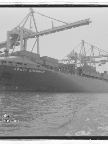 Schwarzweißaufnahme eines großen Containerschiffs mit der Aufschrift CONTI SHANGHAI, am Hafen unter mehreren hohen Ladekränen liegend, mit Wasser im Vordergrund und Museumswasserzeichen unten links.
