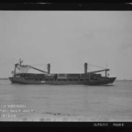 Schwarzweißaufnahme eines Frachtschiffs mit zwei Deckkränen und beladenen Laderäumen, das ruhig auf dem Meer vor einem flachen Horizont fährt; links unten Signet des Internationalen Maritimen Museums Hamburg und Schriftzug ILFORD PAN F am Bildrand.