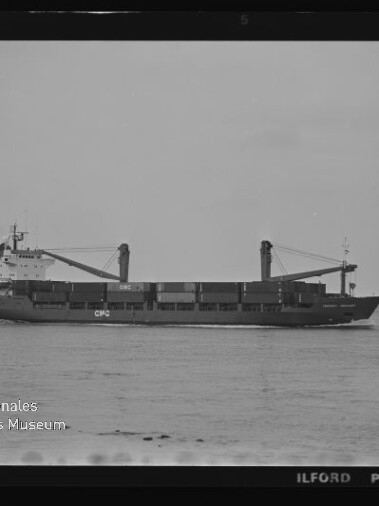 Schwarzweißaufnahme eines Frachtschiffs mit zwei Deckkränen und beladenen Laderäumen, das ruhig auf dem Meer vor einem flachen Horizont fährt; links unten Signet des Internationalen Maritimen Museums Hamburg und Schriftzug ILFORD PAN F am Bildrand.