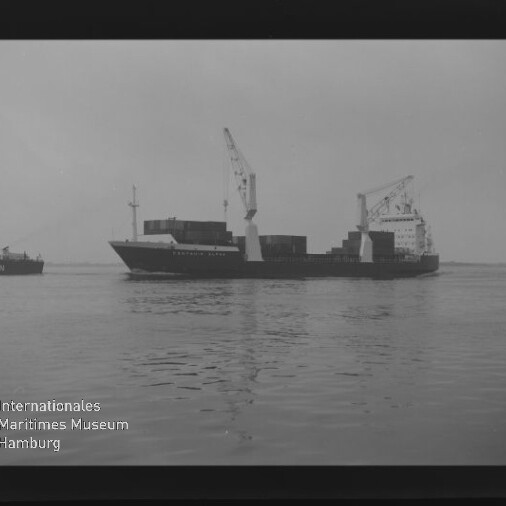 Schwarzweiß-Foto eines Frachtschiffs mit zwei Ladekränen und gestapelten Containern auf ruhigem Wasser, links teilweise ein zweites Schiff sichtbar und unten links das Logo des Internationalen Maritimen Museums Hamburg.