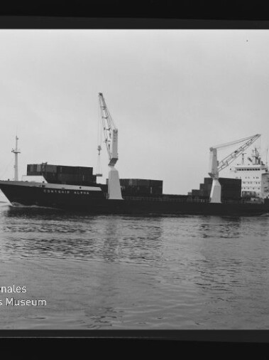 Schwarzweißaufnahme eines beladenen Frachtschiffs mit zwei großen Ladekränen und gestapelten Containern, das ruhig über das Wasser vor flachem Horizont fährt; sichtbarer Filmrand mit 