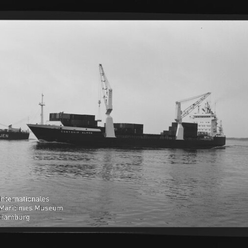 Schwarzweißaufnahme eines beladenen Frachtschiffs mit zwei großen Ladekränen und gestapelten Containern, das ruhig über das Wasser vor flachem Horizont fährt; sichtbarer Filmrand mit 