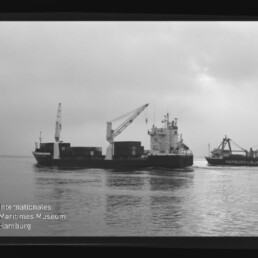 Schwarzweißaufnahme zweier Frachtschiffe auf ruhigem Wasser: links ein containerbeladener Frachter mit Ladekränen, rechts ein kleineres Schiff mit Kran und der Aufschrift ‚Hamburg-Linien‘, vor bedecktem Himmel und mit sichtbarem Museumslogo und Filmrand.