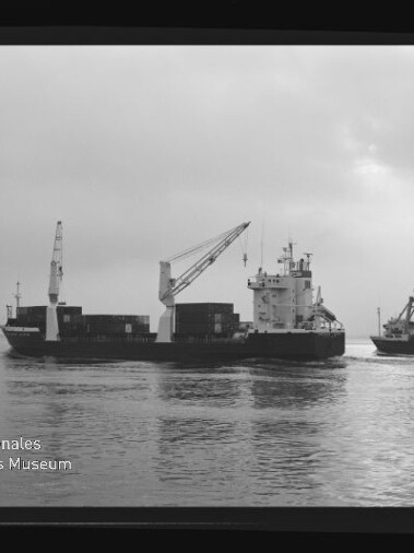 Schwarzweißaufnahme zweier Frachtschiffe auf ruhigem Wasser: links ein containerbeladener Frachter mit Ladekränen, rechts ein kleineres Schiff mit Kran und der Aufschrift ‚Hamburg-Linien‘, vor bedecktem Himmel und mit sichtbarem Museumslogo und Filmrand.