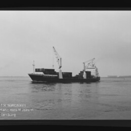 Schwarzweißaufnahme eines Containerschiffs mit zwei Deckkränen und gestapelten Ladecontainern, das bei ruhigem Wasser unter grauem Himmel fährt; unten links sichtbar das Logo und der Schriftzug des Internationalen Maritimen Museums Hamburg.
