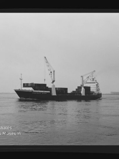 Schwarzweißaufnahme eines Containerschiffs mit zwei Deckkränen und gestapelten Ladecontainern, das bei ruhigem Wasser unter grauem Himmel fährt; unten links sichtbar das Logo und der Schriftzug des Internationalen Maritimen Museums Hamburg.