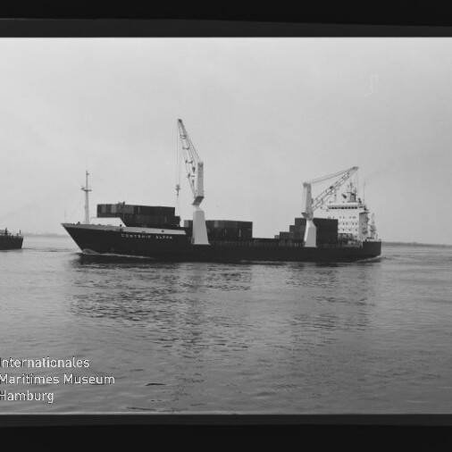 Schwarzweißaufnahme eines Containerfrachters mit zwei Bordkränen und gestapelten Containern, fahrend auf ruhigem Wasser; links ein weiteres Schiff am Bildrand und unten links das Logo des Internationalen Maritimen Museums Hamburg, sichtbar innerhalb des Filmrands.