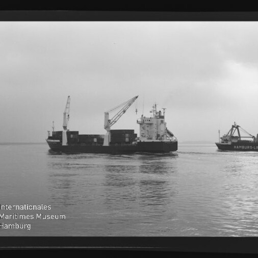 Schwarzweiß-Aufnahme eines Frachtschiffs mit ausfahrbaren Ladekränen und Containerstapel, rechts ein weiteres Schiff mit dem Schriftzug HAMBURGLINJEN, auf ruhiger See unter trübem Himmel; unten links Logo und Schriftzug des Internationalen Maritimen Museums Hamburg.