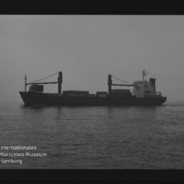 Schwarz-weiß-Aufnahme eines Frachtschiffs mit zwei Ladekränen und markantem Decksaufbau, das ruhig auf dem Meer vor einem nebligen Horizont liegt; unten links das Logo des Internationalen Maritimen Museums Hamburg sichtbar.