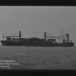 Seitenansicht eines mit Containern beladenen Frachtschiffs mit zwei Ladekränen auf ruhiger See, Schwarzweiß-Foto mit Filmrand und Logo des Internationalen Maritimen Museums Hamburg.
