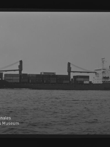 Seitenansicht eines mit Containern beladenen Frachtschiffs mit zwei Ladekränen auf ruhiger See, Schwarzweiß-Foto mit Filmrand und Logo des Internationalen Maritimen Museums Hamburg.