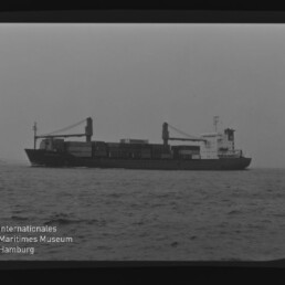 Schwarzweiß-Foto eines Containerschiffs in Seitenansicht auf ruhiger See mit gestapelten Containern, zwei Ladekränen und dem Logo des Internationalen Maritimen Museums Hamburg in der unteren linken Ecke.