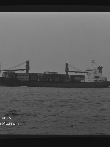 Schwarzweiß-Foto eines Containerschiffs in Seitenansicht auf ruhiger See mit gestapelten Containern, zwei Ladekränen und dem Logo des Internationalen Maritimen Museums Hamburg in der unteren linken Ecke.