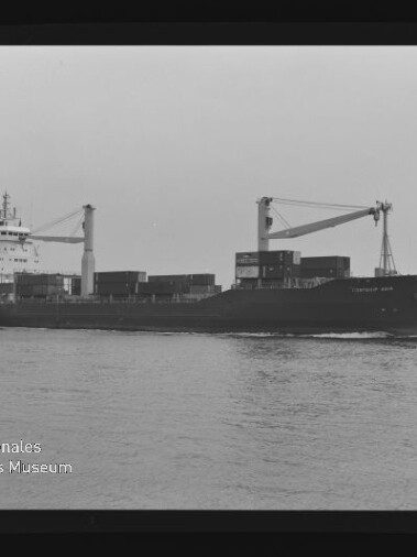 Schwarzweißaufnahme eines Containerfrachters mit gestapelten Containern und zwei Auslegerkränen, der über ruhiges Wasser fährt; unten links ist das Logo des Internationalen Maritimen Museums Hamburg zu sehen.