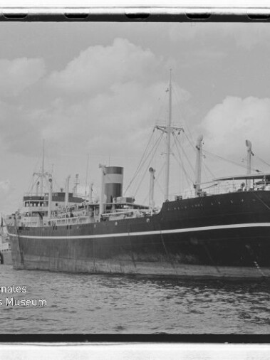 Großes Frachtschiff „Alisios Bremen“ längsseits im Hafen, Seitenansicht mit markantem Schornstein, Ladekranen und Flagge am Heck vor bewölktem Himmel.