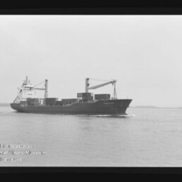 Containerschiff mit zwei Deckskranen und gestapelten Ladecontainern fährt ruhig auf dem Meer; unten links sichtbar das Logo des Internationalen Maritimen Museums Hamburg.