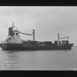Schwarz-weiß-Seitenansicht eines Frachtschiffs mit zwei Ladekränen und gestapelten Containern auf ruhigem Wasser unter bedecktem Himmel; Bild zeigt Filmrand mit AGFA APX 100-Markierung und Wasserzeichen des Internationalen Maritimen Museums Hamburg.